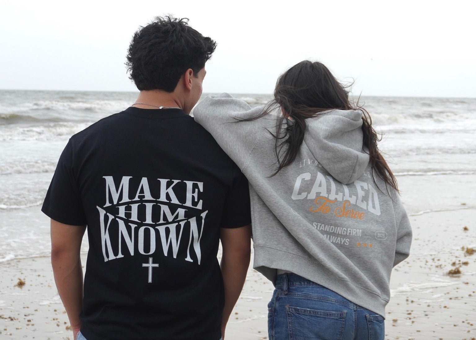 Two people standing on a beach, facing away from the camera, wearing Make Him Known t-shirt and Called To Serve Hoodie.